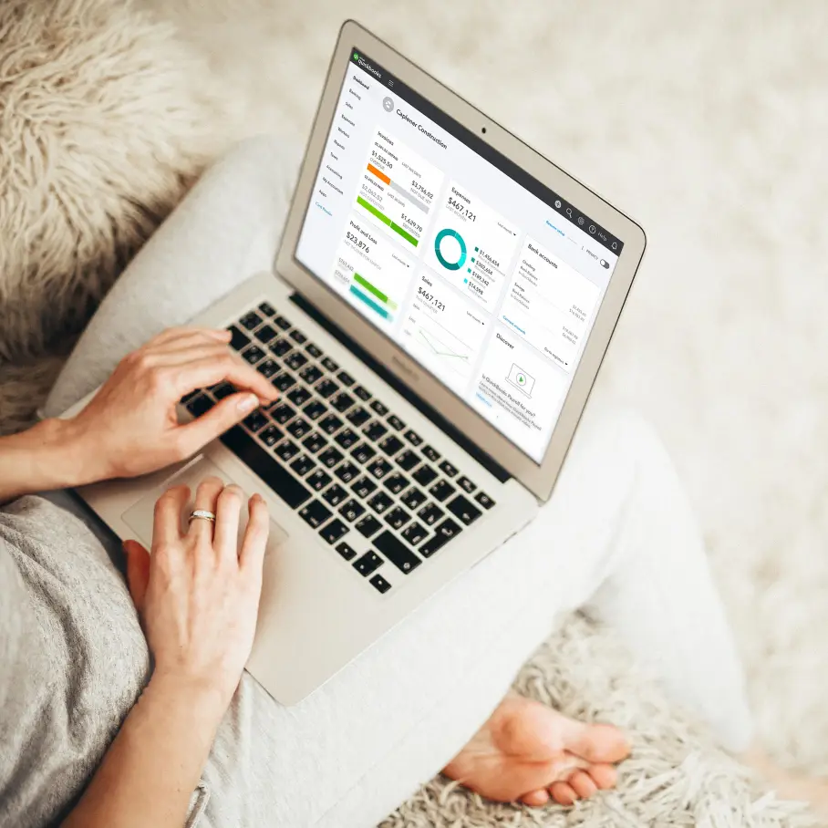 A woman working on a laptop while sitting on a cozy, textured rug, managing business finances through the Quickbooks online dashboard